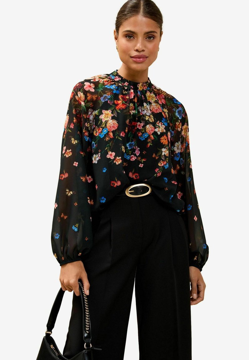 Blusa nera con fantasia floreale caratterizzata da colorati motivi di fiori, scollatura arricciata e maniche lunghe a palloncino. Abbinata a pantaloni neri a vita alta e una borsa.