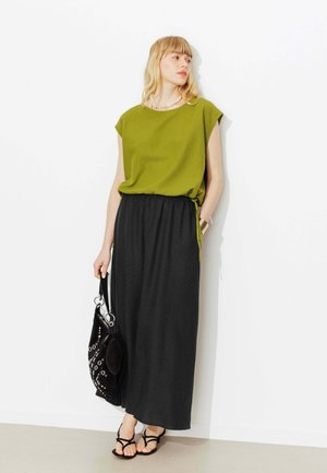 Femme portant un haut vert sans manches, une longue jupe noire, des sandales noires à lanières, tenant un sac à main noir avec des œillets métalliques, debout contre un mur blanc.