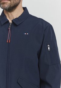 Navy blue jacket featuring a zipped front, side pockets, and a small multicolor logo on the chest. Textured, sturdy fabric with a collar.