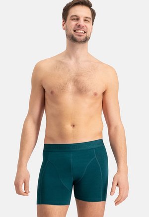 Uomo sorridente che indossa boxer verde acqua con elastico in vita con la scritta "Bamboo basics", in piedi contro uno sfondo chiaro uniforme, con le braccia rilassate lungo i fianchi.