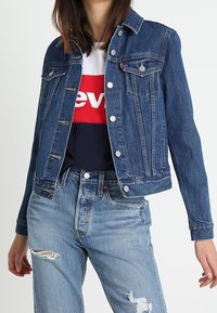 Woman wearing a buttoned blue denim jacket over a red, white, and navy logo t-shirt with ripped light blue jeans.
