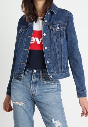 Frau trägt eine knöpfige blaue Jeansjacke über einem rot-weißen und marineblauen Logo-T-Shirt mit zerrissenen hellblauen Jeans.