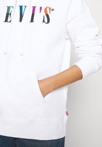 Sudadera blanca con el logo "LEVI'S" multicolor, bolsillo canguro en el frente y cordones, combinada con jeans azules.