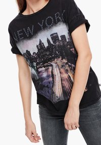 Camiseta gráfica negra con la palabra "NEW YORK" en tachuelas, con un diseño impreso de un horizonte urbano y la Estatua de la Libertad. Manga corta, corte relajado.