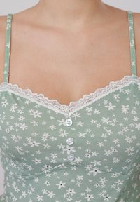 Femme portant un caraco vert à motifs floraux avec une bordure en dentelle blanche et trois boutons sur le devant, montrant le haut de la poitrine et les épaules.