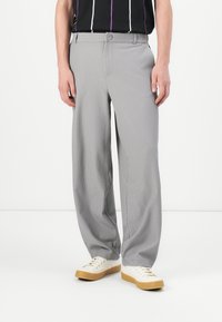 Mand iført lysegrå plisserede bukser, hvide sneakers med gummisåler og en sort skjorte med lodrette hvide og lilla striber.