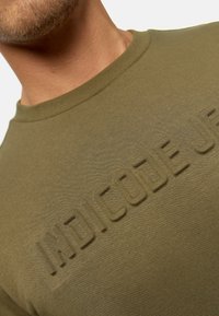 Camiseta de manga corta en color verde oliva, hecha de una tela suave, con el texto en relieve "INDICODE" en el frente y un diseño de cuello redondo.