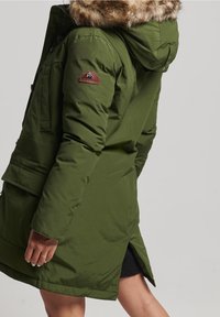 Persoon draagt een olijfgroene parka met een schapenvacht gevoerde capuchon en een merkembleem op de bovenarm, die tot onder de midden dij reikt.