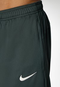 Nike treningsshorts i mørkegrønn stoff, med elastisk linning, sidelommer og en hvit logo nederst til venstre.