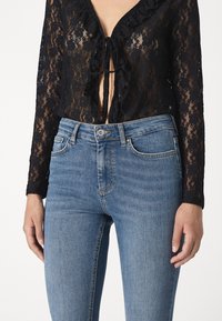 Blusa de encaje negro con cuello con volantes y detalle de lazo, combinada con jeans de denim azul claro que cuentan con bolsillos clásicos y una silueta ajustada.