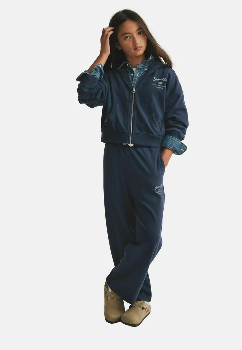 Felpa con zip blu navy con tasche frontali, indossata sopra una camicia di jeans, abbinata a pantaloni della tuta blu navy coordinati e zoccoli beige. Dettagli di marchio semplici.