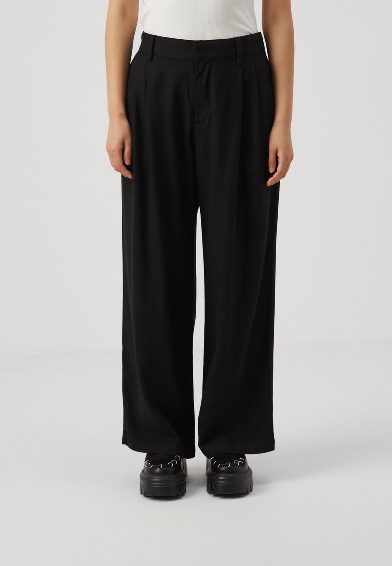 Svarta vida byxor med framfåror, tillverkade av lättviktsmaterial. Har en skräddarsydd midja och matchas med kraftiga svarta plattformsskor.