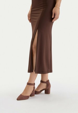 Kobieta w brązowej midi spódnicy z przednim rozcięciem i dopasowanych brązowych zamszowych sandałach na słupku z paskiem wokół kostki, stojąca na białym tle.