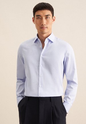 Camicia azzurro chiaro con colletto aperto, maniche lunghe e una texture sottile. Indossata con pantaloni scuri su misura, che mettono in evidenza un design aderente.
