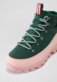 Bota de senderismo verde con parte superior de tela texturizada, suela de goma rosa, cordones reflectantes y un tirador etiquetado como "HUNTER" en rojo.