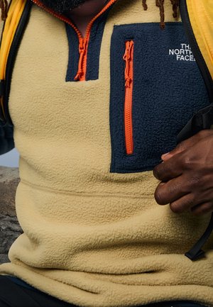 Fleece pullover in beige met marineblauwe accenten. Voorzien van een oranje rits en borstzak. Glad textuur met een ontspannen pasvorm.