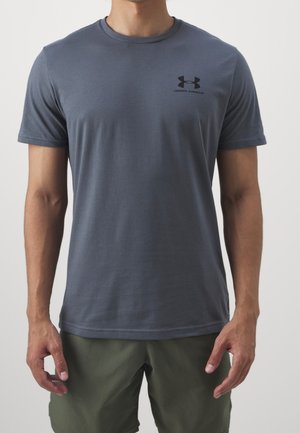 Mężczyzna w krótkim rękawku ciemnoszarej koszulce Under Armour i oliwkowo-zielonych szortach stoi na jednolitym tle.