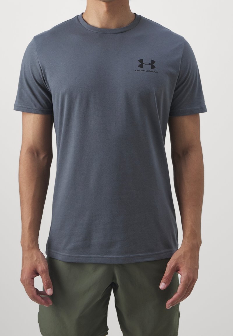Homme portant un t-shirt à manches courtes gris foncé Under Armour et un short vert olive, debout devant un fond uni.