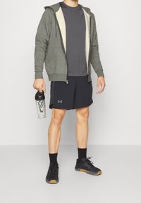 Felpa grigia con zip e fodera leggera, t-shirt grigia scura, pantaloni corti sportivi neri e scarpe da ginnastica nere. Tiene in mano una bottiglia shaker trasparente.