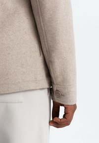 Beige Wolljacke mit strukturierter Oberfläche, ausgestattet mit einer Knopfleiste am Ärmel und einer Seitentasche, getragen über hellen Hosen.