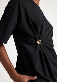 T-shirt in cotone nero con design intrecciato e un bottone centrale dorato come accento. Maniche corte e vestibilità comoda con texture liscia.