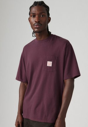 Camiseta de manga corta color marrón, hecha de algodón. Presenta un bolsillo en el pecho con una pequeña etiqueta roja y blanca. Diseño clásico de cuello redondo.