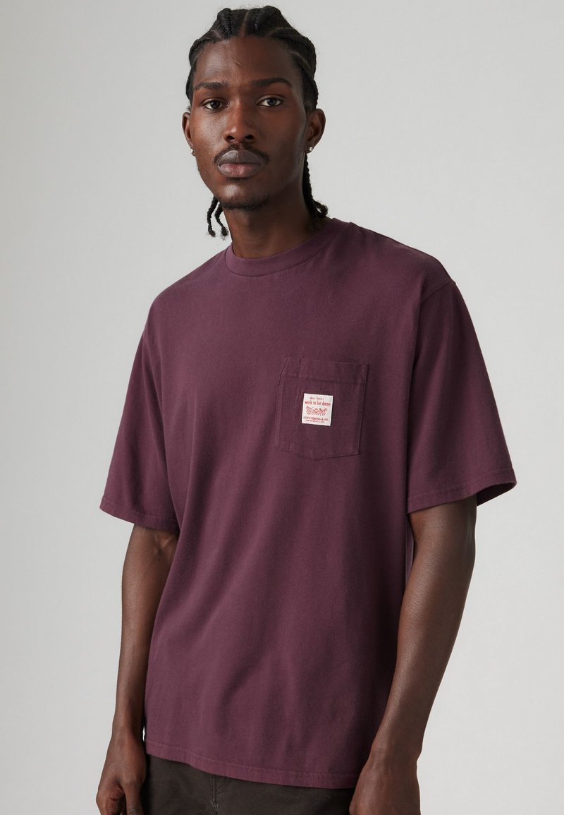 Bordeaux kleurig T-shirt met korte mouwen van katoen. Voorzien van een voorkeurszak op de borst met een klein rood en wit label. Klassiek rond decolleté ontwerp.
