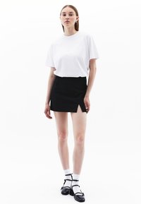 Witte T-shirt gemaakt van katoen, gecombineerd met een zwarte minirok met een zijsplit. Model draagt witte sokken en zwarte sandalen met bandjes.