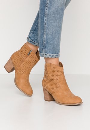 Brune ankelstøvle lavet af ruskind med perforeret design, lynlås lukning og en stablet hæl. Bæres med cropped blå jeans.
