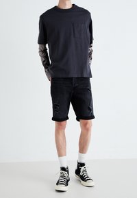 Camiseta negra de algodón con un bolsillo en el pecho, llevada sobre una camiseta de manga larga con estampado, combinada con pantalones cortos de mezclilla negros desgastados y zapatillas negras.