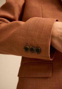 Tejido texturizado marrón con un diseño entallado. Tres botones oscuros son visibles en el puño de la prenda. Líneas simples y limpias con forma estructurada.
