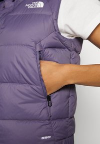 Fioletowa pikowana kamizelka z kieszenią na zamek, z przeszywanym wzorem i białym logo. Tkanina jest gładka i lekka, zaprojektowana do noszenia warstwowego.