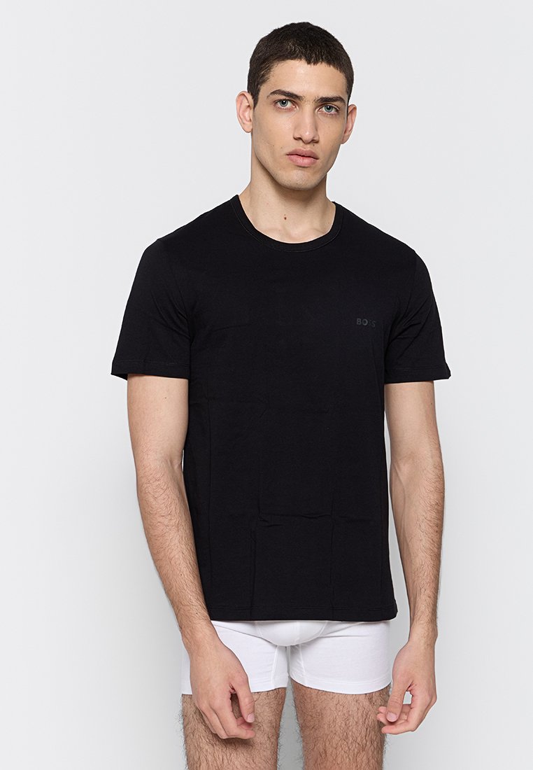 Jonge man met kort donker haar, gekleed in een eenvoudig zwart T-shirt met korte mouwen en witte boxershorts, staande tegen een lichte achtergrond.