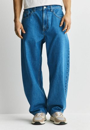 Muž v pohodlných modrých džínsach a béžových teniskách, stojaci pred jednoduchým svetlým pozadím.