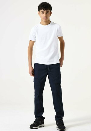 Teenager with curly hair wearing a plain white t-shirt, navy cargo pants, and black sneakers, standing against a plain white background.