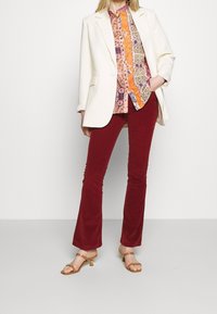 Blazer blanc porté sur un chemisier coloré à motifs orange, rose et bleu. Pantalon évasé bordeaux et talons à brides beige complètent la tenue.