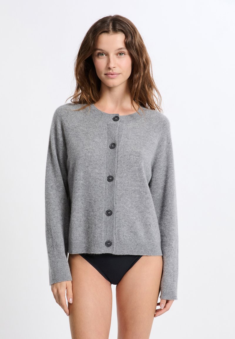 Jeune femme avec des cheveux bruns ondulés portant un cardigan gris boutonné et un sous-vêtement noir, se tenant devant un fond blanc uni.