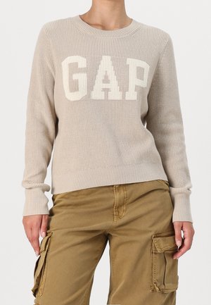 Beige striktrøje med store hvide "GAP" bogstaver kombineret med lysebrune cargobukser med sidelommer vist fra hals til hofter.