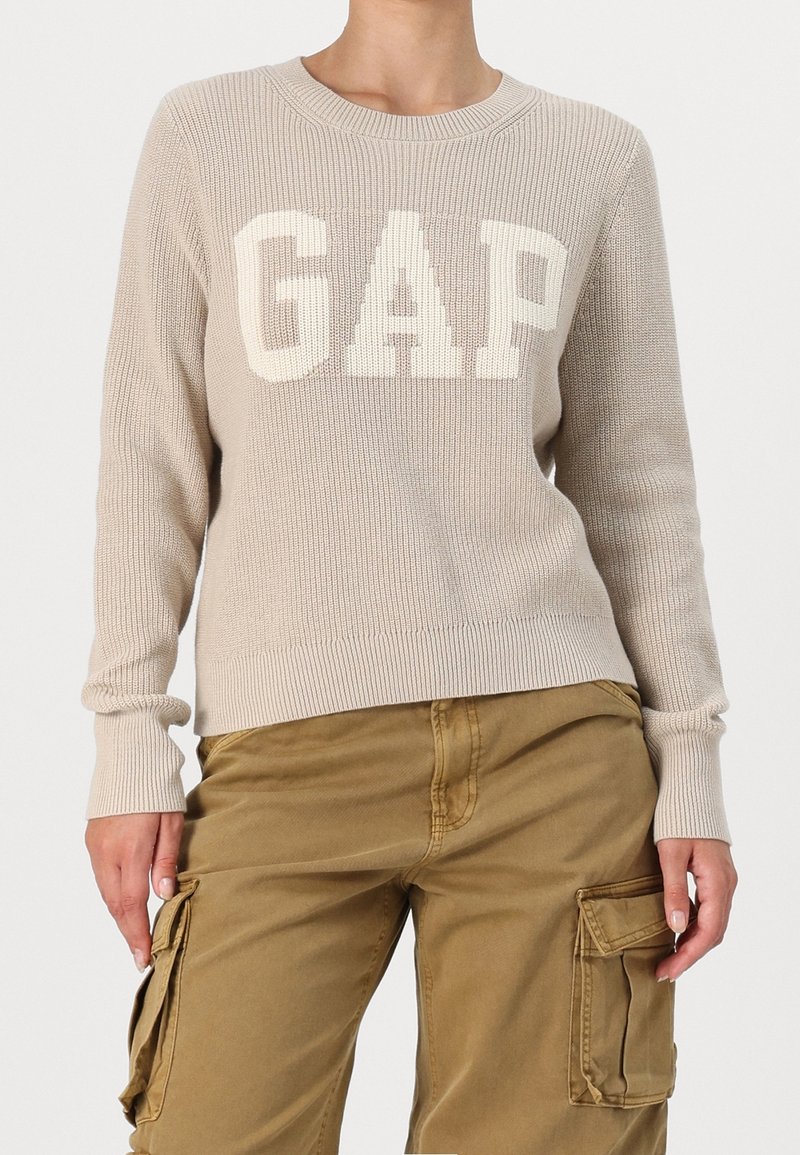Pull en maille beige avec de grandes lettres blanches "GAP" associé à un pantalon cargo marron clair avec poches latérales, présenté du cou jusqu'aux hanches.
