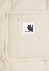 Bežinė pūkinė striukė su siuvinėta tekstūra, turinti ryškų kvadratinį pleistrą su užrašu "Carhartt" ir juodu logotipu.