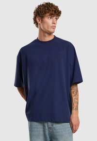Camiseta oversized de color azul marino en tejido suave, con cuello redondo y hombros caídos. Tatuajes coloridos visibles en el antebrazo.
