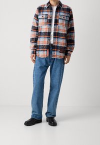 Červeno-černá a modrá károvaná flanelová košile s dvěma kapsami na prsou, nošená přes bílý tričko. V páru s volnými modrými džíny a černými botami.