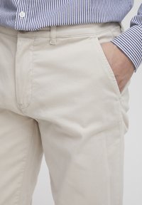 Beige Baumwollhosen mit Reißverschluss- und Knopfverschluss, Seitentaschen und einer glatten Textur. Getragen mit einem blau-weißen gestreiften Hemd.