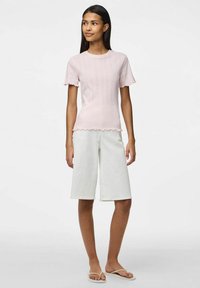 Jasnoróżowa, prążkowana koszulka z krótkimi, falbankowymi rękawami, połączona z białymi, dżinsowymi szortami do kolan. Modelka nosi klapki, stoi w zaawansowanej pozycji.