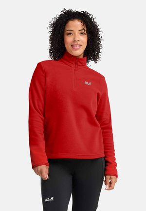 Vrouw met krullend haar, gekleed in een rood fleecevest met een halve rits en een zwarte broek met logo's, staand en glimlachend tegen een effen achtergrond.