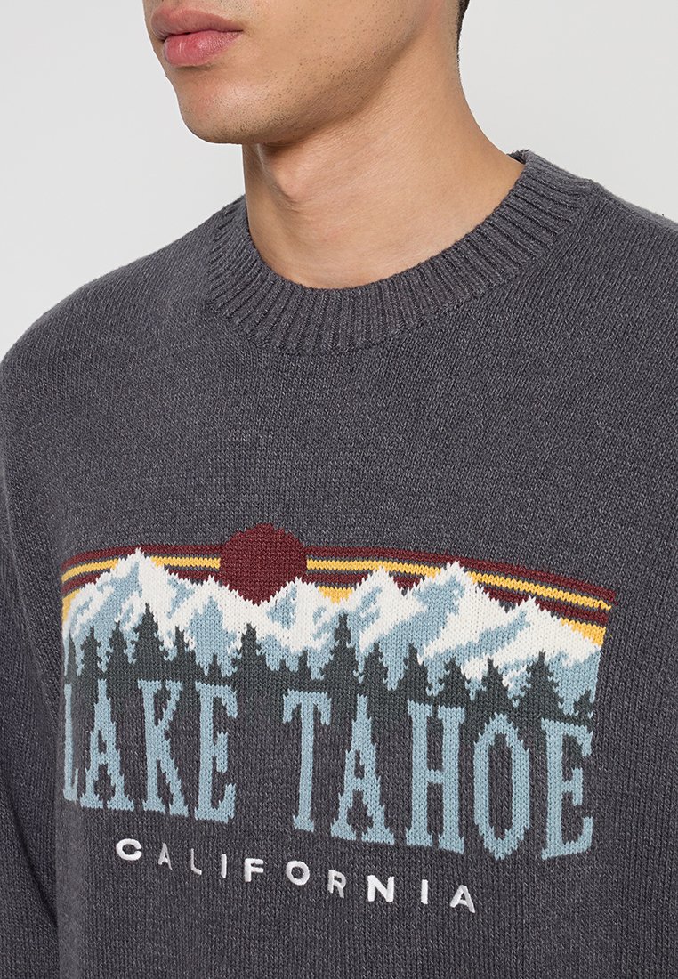 Grauer gestrickter Pullover mit Grafik von einem Berg und der Sonne. Text lautet "LAKE TAHOE CALIFORNIA" in kontrastierenden Farben. Gerippter Kragen.