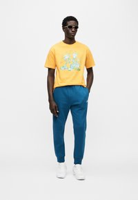 Κίτρινη βαμβακερή μπλούζα με καρτούν γραφικό; πετρόλ παντελόνι φόρμας; λευκά αθλητικά παπούτσια; γυαλιά ηλίου. Απλός σχεδιασμός, casual εφαρμογή, χωρίς ορατά σχέδια.