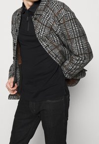 Šedá a hnědá kostkovaná bunda s velkými knoflíky, s límečkem a texturovanou tkaninou, nošená přes černou košili a tmavé džíny.