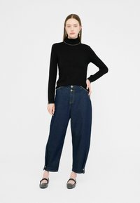 Zwarte coltrui, gecombineerd met loszittende donkerblauwe denim jeans met zijknoppen en een onafgebroken zoom. Eenvoudige platte zwarte schoenen.