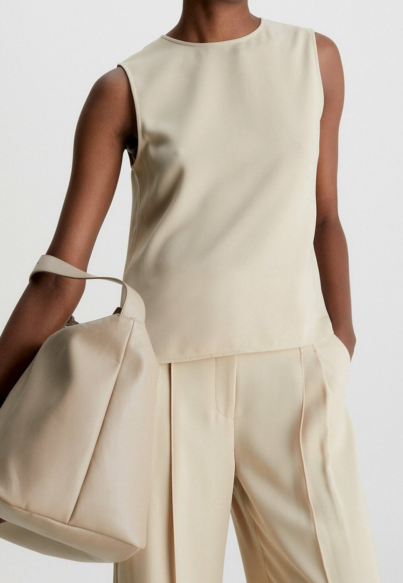 Beige ärmlös topp med en mjuk textur, kombinerad med matchande byxor och en stor, mjuk väska över armen.